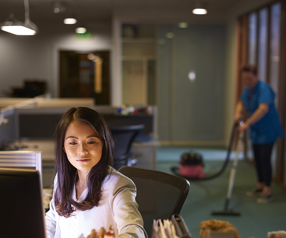 office worker behind her laptop, cleaning lady hovering in the background